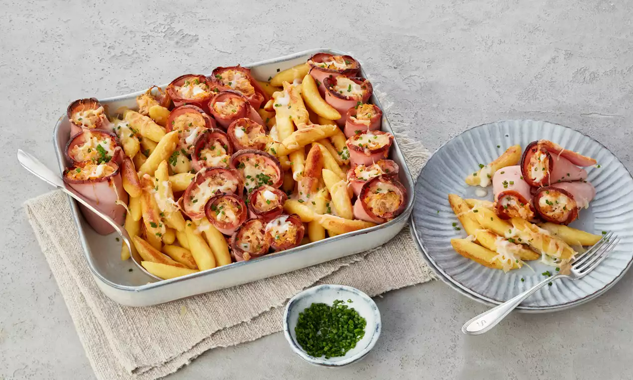 Goldbraune Penne und mit Käse gefüllte Leberkäseröllchen, gebacken in einer silbernen Auflaufform, garniert mit frischen Schnittlauchringen