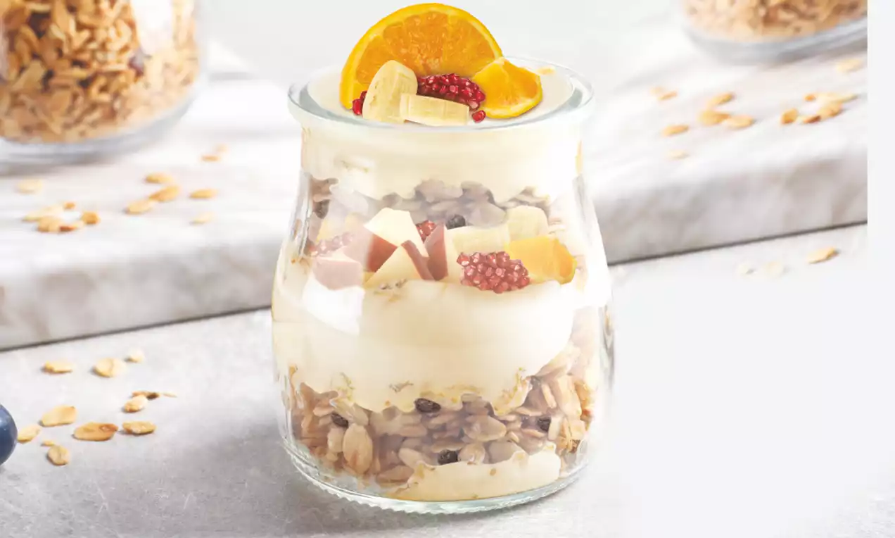 Creamy pale pudding layered with golden-brown granola in a clear glass jar, topped with orange slice, banana, berries, and granola