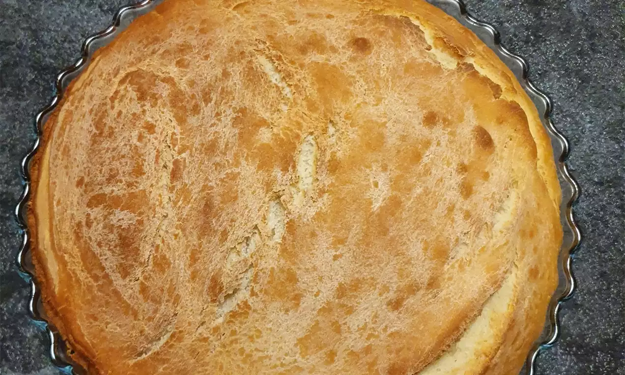 Okrugli kruh zlatnosmeđe boje s lagano ispucalom korom u valovitoj metalnoj posudi na tamnoj, grubi podlozi