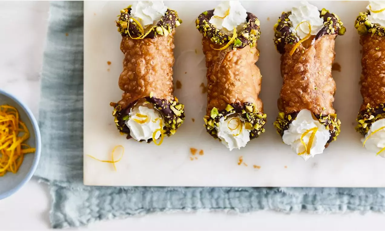 Хрупкави цилиндри с топъл карамелен цвят, краища с черен шоколад и ядки, пълнеж от бяла сметана и портокалови стърготини върху мрамор