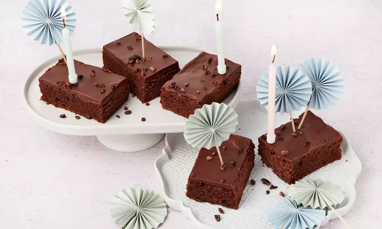 Schokoladenschnitten mit dunkler Glasur und Kakaostückchen auf weißen Platten, verziert mit Kerzen und Papierfächern in Pastellfarben