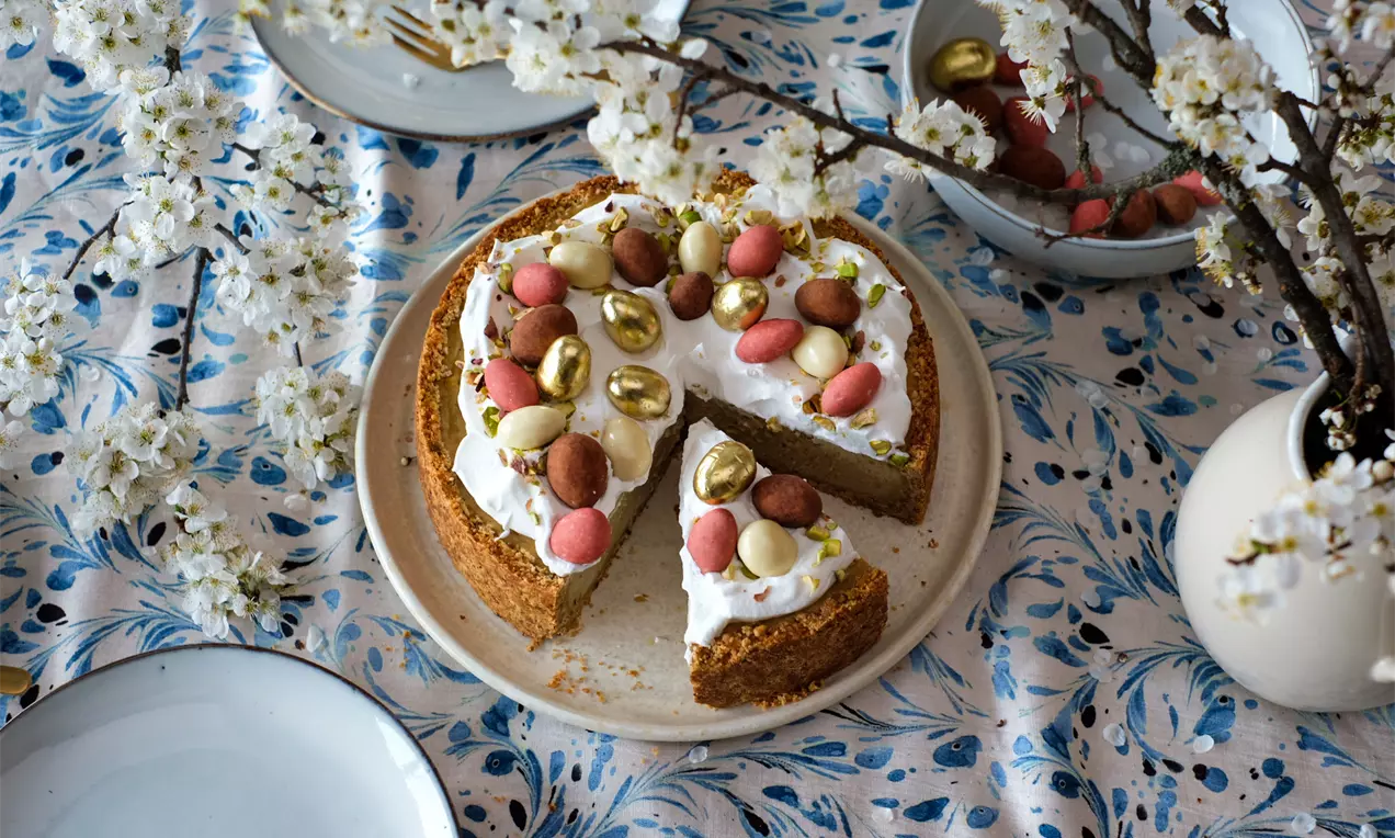 Чийзкейк с шамфъстък с бледозелен крем върху кафяв блат, покрит с бяла сметана и декориран с цветни шоколадови яйчица