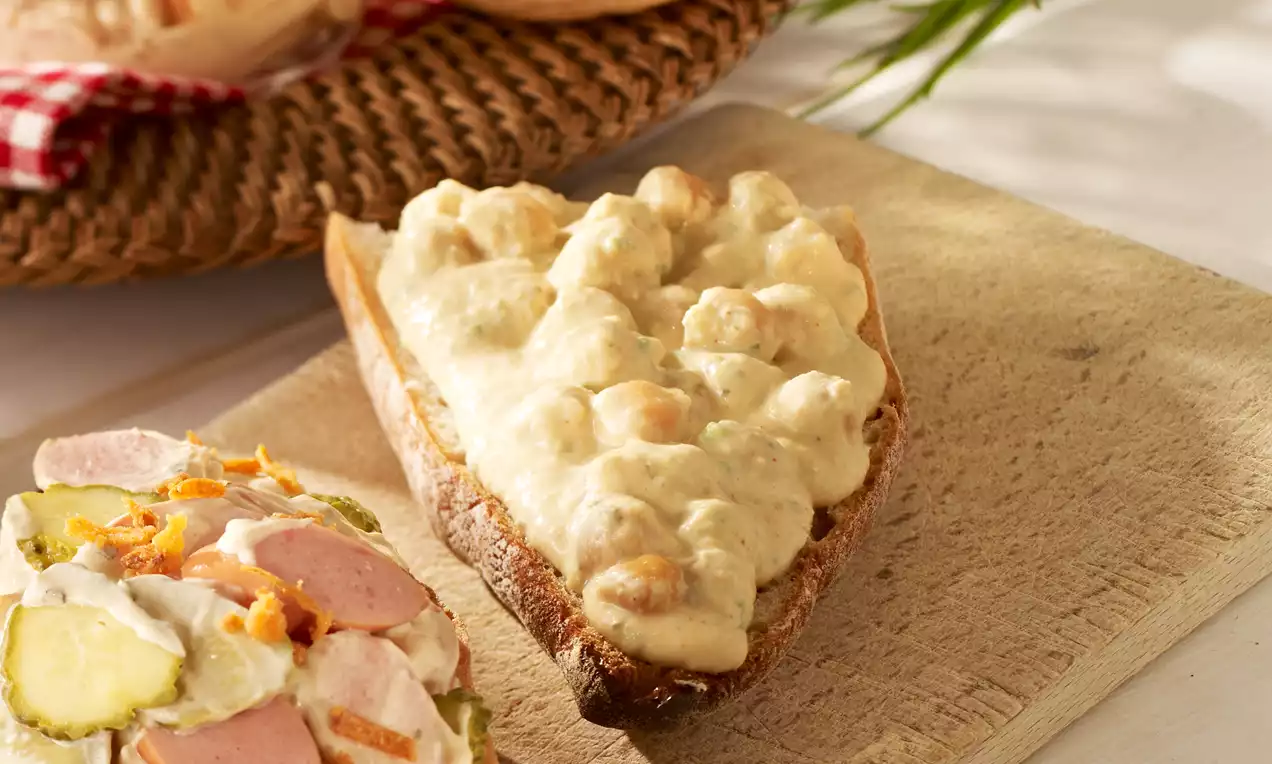 Cremige, beige Kichererbsencreme mit weicher Textur auf einer rustikalen Scheibe dunklem Brot, serviert auf einem hölzernen Brett