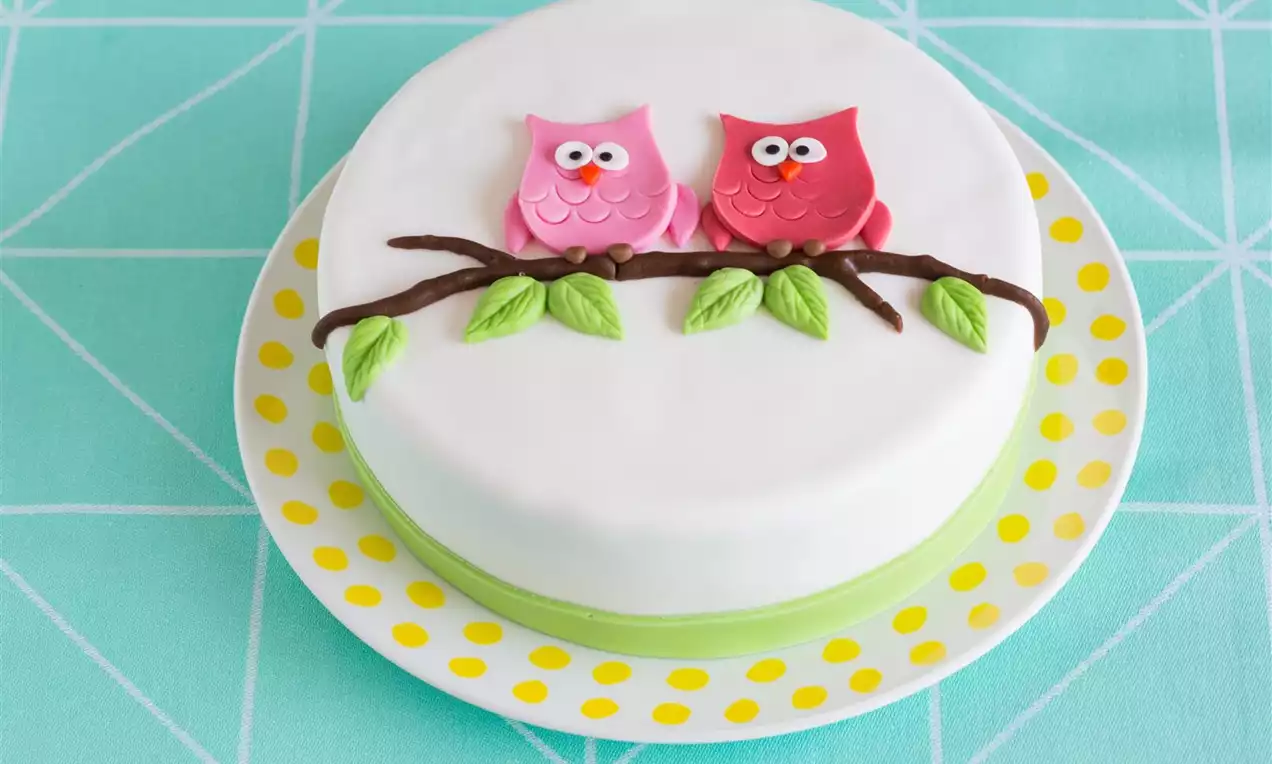 Rund kake med hvit glasur, rosa og røde ugler i fondant på brun grein med grønne blader, plassert på gulprikket fat mot turkis duk