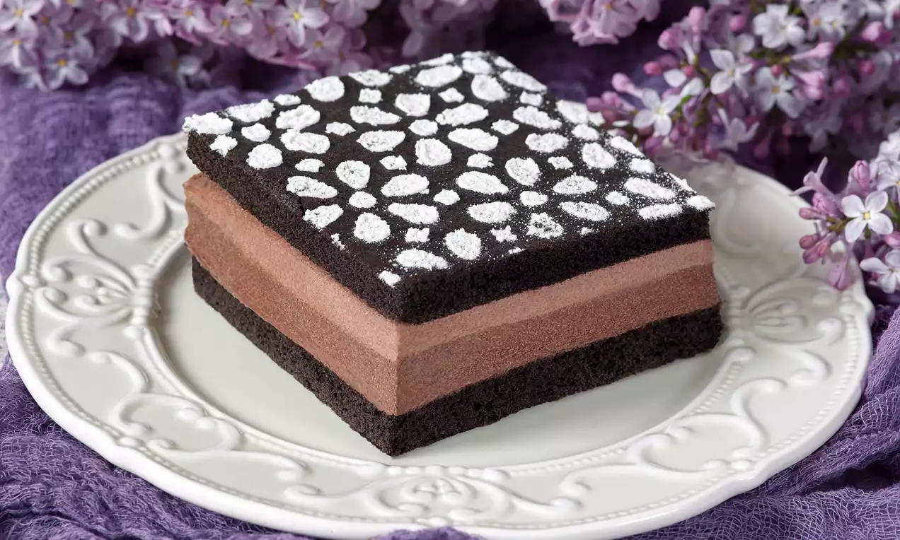 Čtvercový dezert s kakaovou vrstvou, čokoládovým krémem a lehkým vzorem z moučkového cukru na bílém talíři, obklopený fialovými květy
