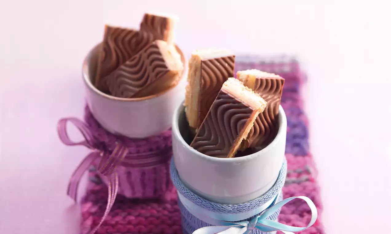 Karamell-Shortbread mit wellenförmiger Schokoladenschicht und hellbeigem Keksboden in weißen Bechern mit farbigen Schleifen arrangiert