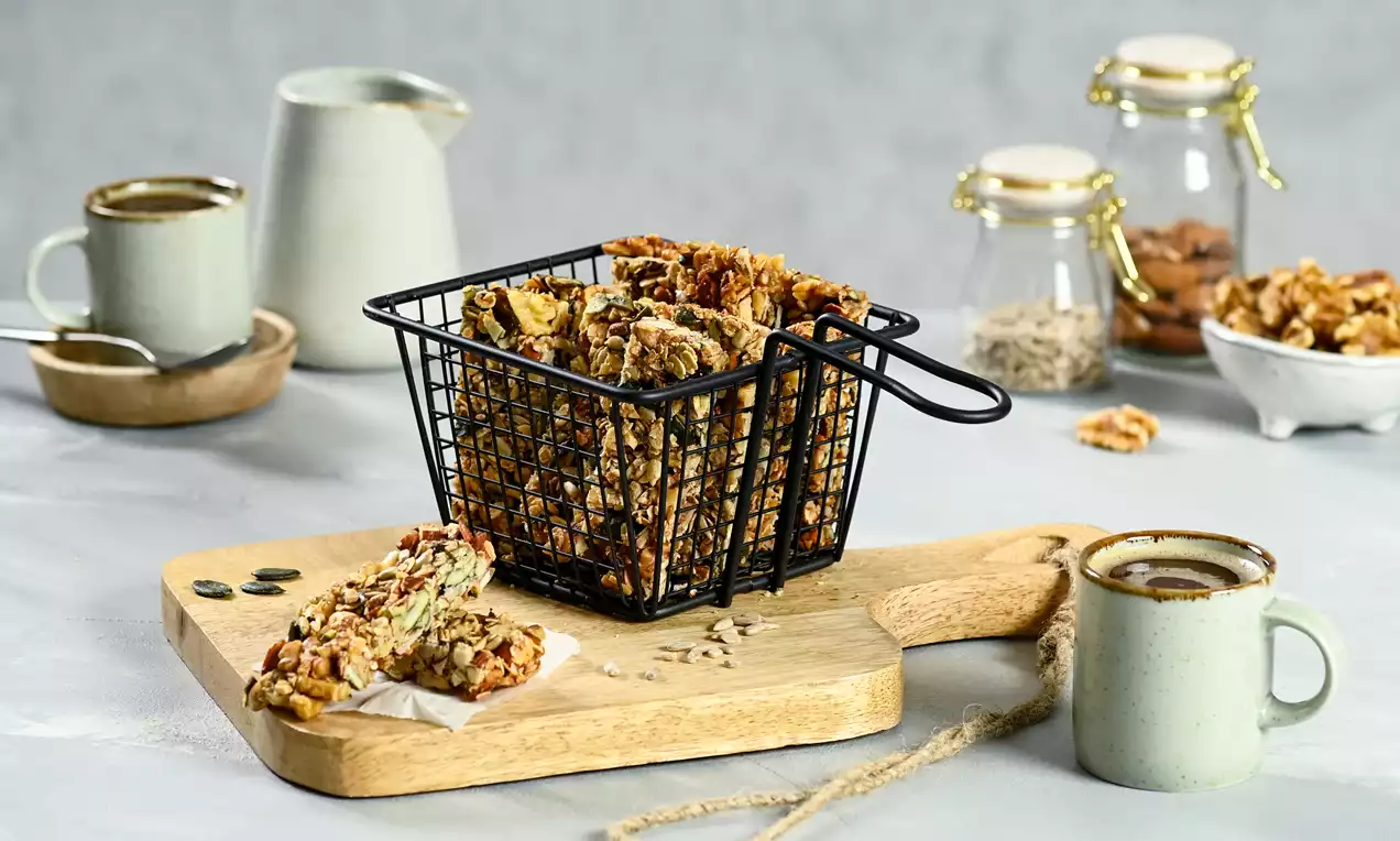 Rustic walnut and seed breakfast bars in a black wire basket on a wooden board, surrounded by jars, mugs, and scattered ingredients
