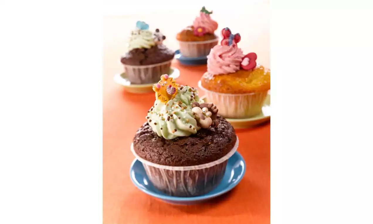 Sjokolademuffins med lysegrønn krem, sjokoladestrø og små sukkerblomster på blå asjett, omgitt av flere fargerike muffins