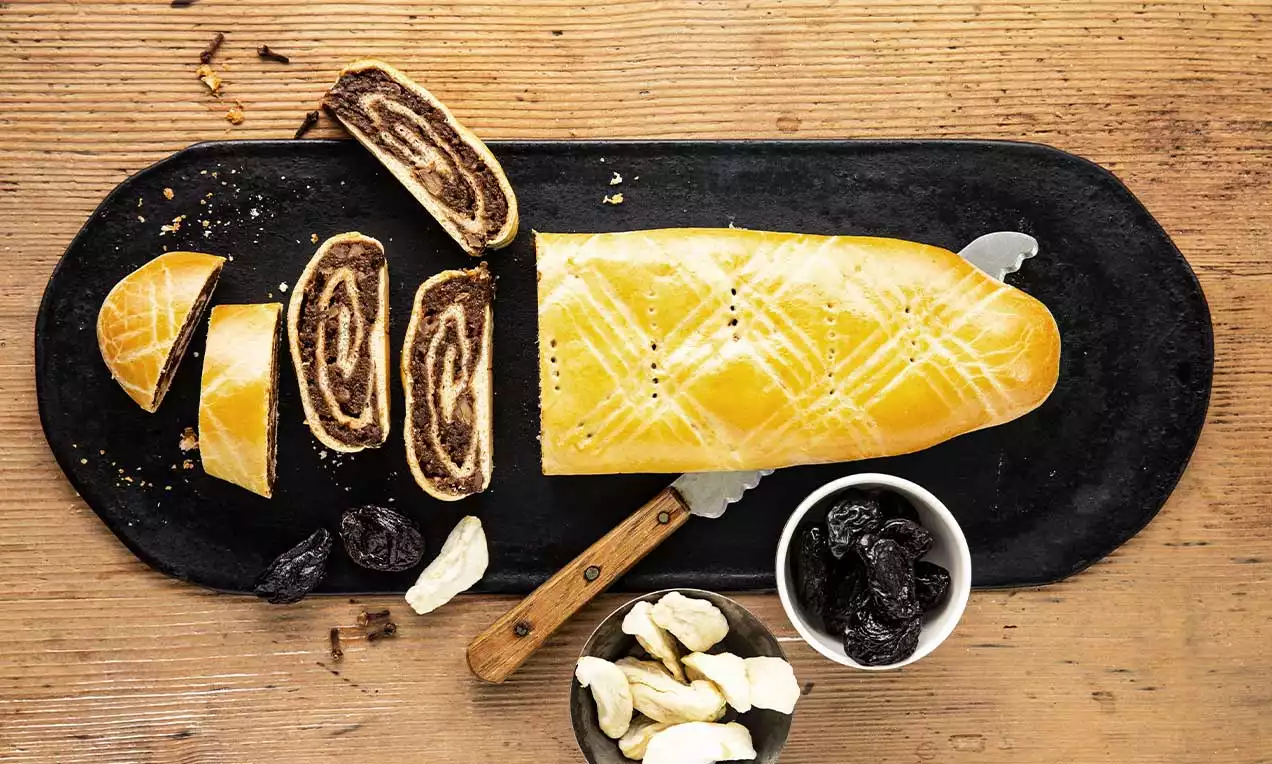 Goldbraune Birewegge mit rautenförmigem Muster, in Scheiben geschnitten auf schwarzem Brett, daneben Trockenfrüchte und Messer