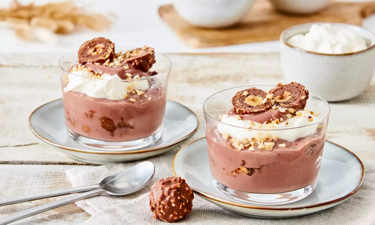 Zwei gläserne Dessertschalen mit cremiger Schokomousse, Sahnehäubchen, gehackten Haselnüssen und Schokopralinen auf einem grauen Teller
