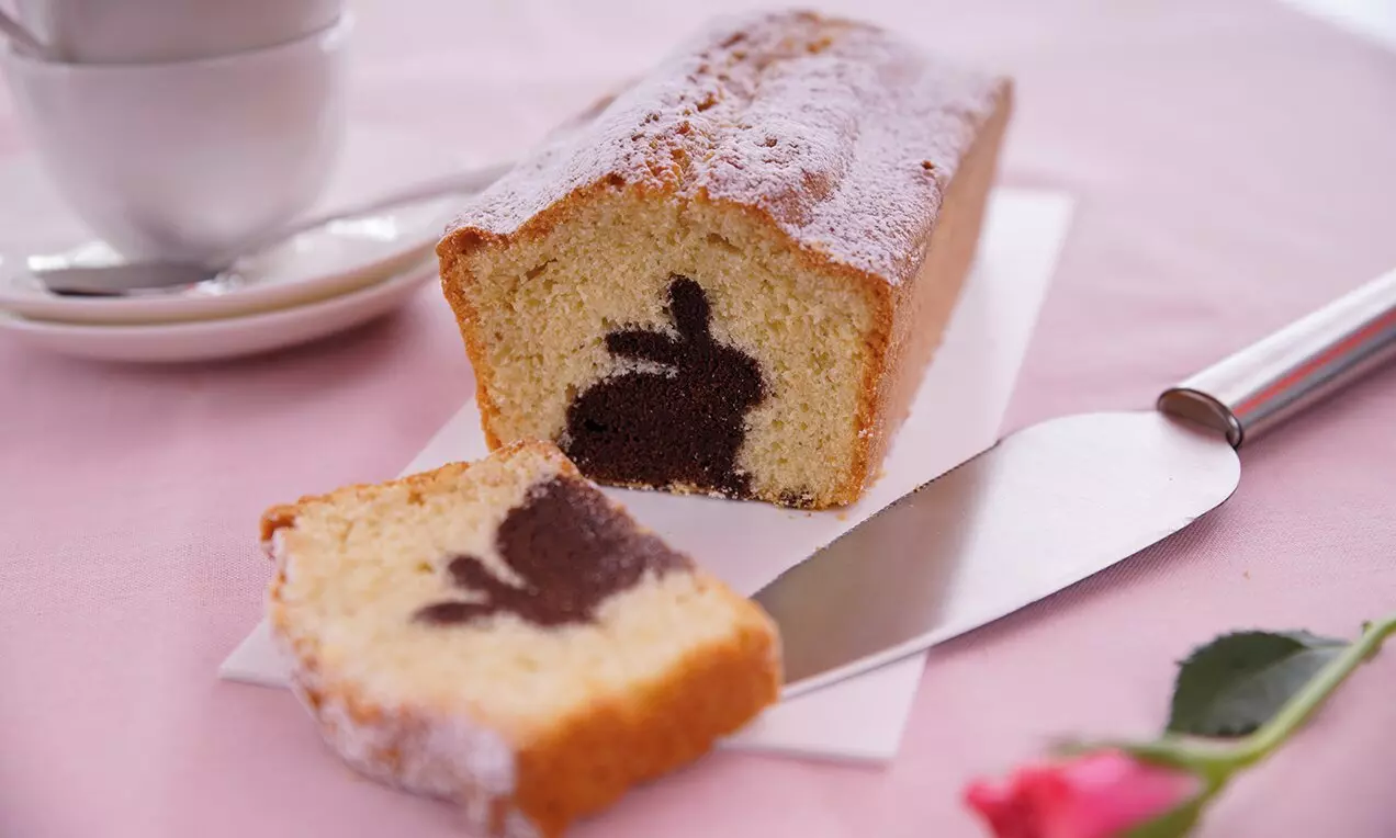 Правоъгълен светложълт кекс с кафява фигура на зайче в разреза, поръсен с пудра захар, върху бял картон и нож до роза и чаша