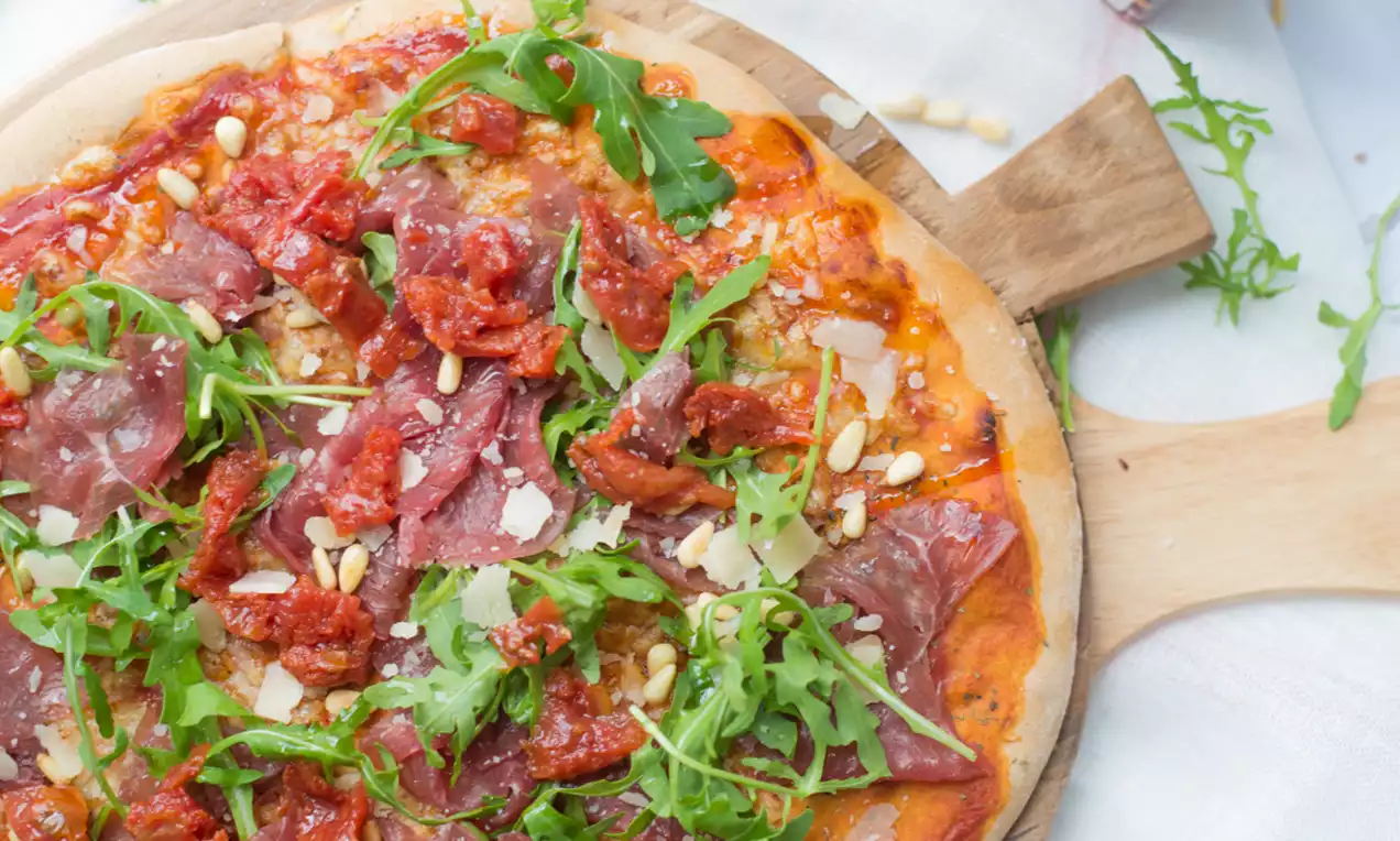 Dunne ronde pizza met een krokante korst, helderrode tomatensaus, flinterdunne carpaccio, rucola, pijnboompitten en geraspte Parmezaan op een houten plank