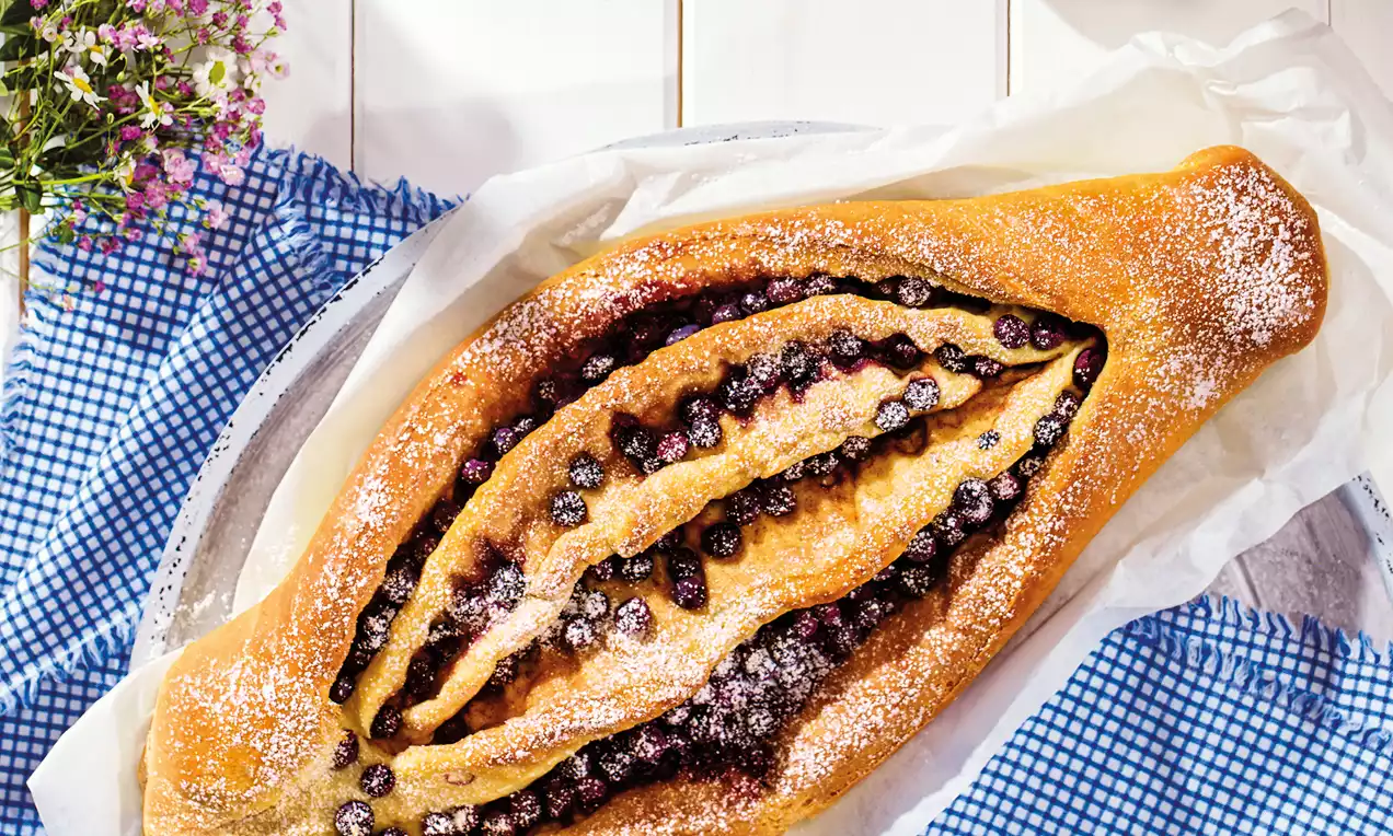 Izduženi pečeni kolač zlatne boje ispunjen sočnim crnim borovnicama, posut šećerom u prahu, na bijelom tanjiru s plavim stolnjakom