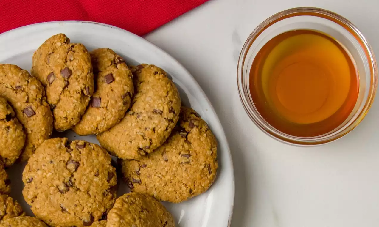 Galletas doradas con textura rústica y trozos de chocolate, dispuestas en círculo sobre un plato blanco junto a un vaso de miel ámbar