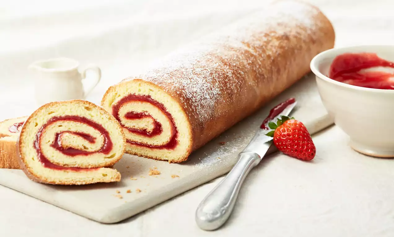 Zarte Biskuitrolle mit hellbrauner Kruste und roter Füllung, zwei Scheiben aufgeschnitten, garniert mit Erdbeeren und Puderzucker