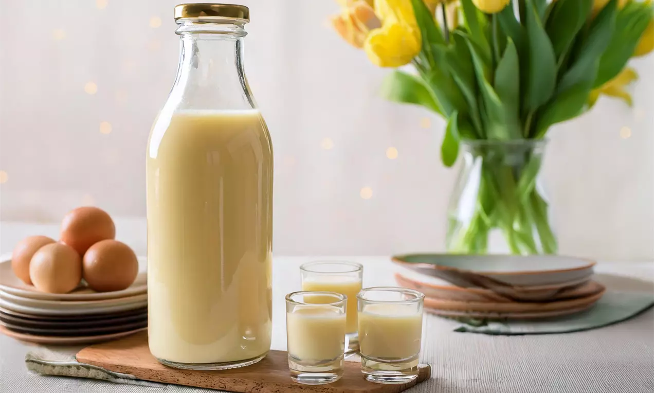 Bouteille en verre transparente remplie de liqueur jaune pâle, entourée de verres assortis sur une planche en bois avec des œufs et des tulipes jaunes
