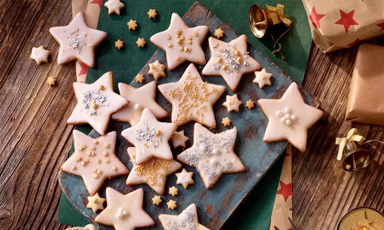 Marcipánové hvězdičky světle hnědé s bílou polevou, zdobené perličkami a třpytem, rozložené na dřevěném podnosu vedle drobných ozdob