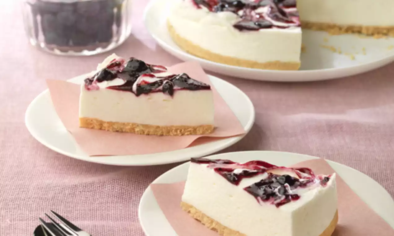Porciones de tarta de yogur blanco sobre base dorada de galleta, con remolinos brillantes de mermelada de arándanos en platos blancos