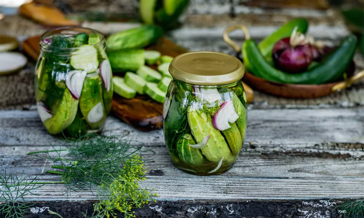 Стъклен буркан с поставени цели зелени краставички, крехки резени червен лук и свежи стръкове копър върху дървена маса