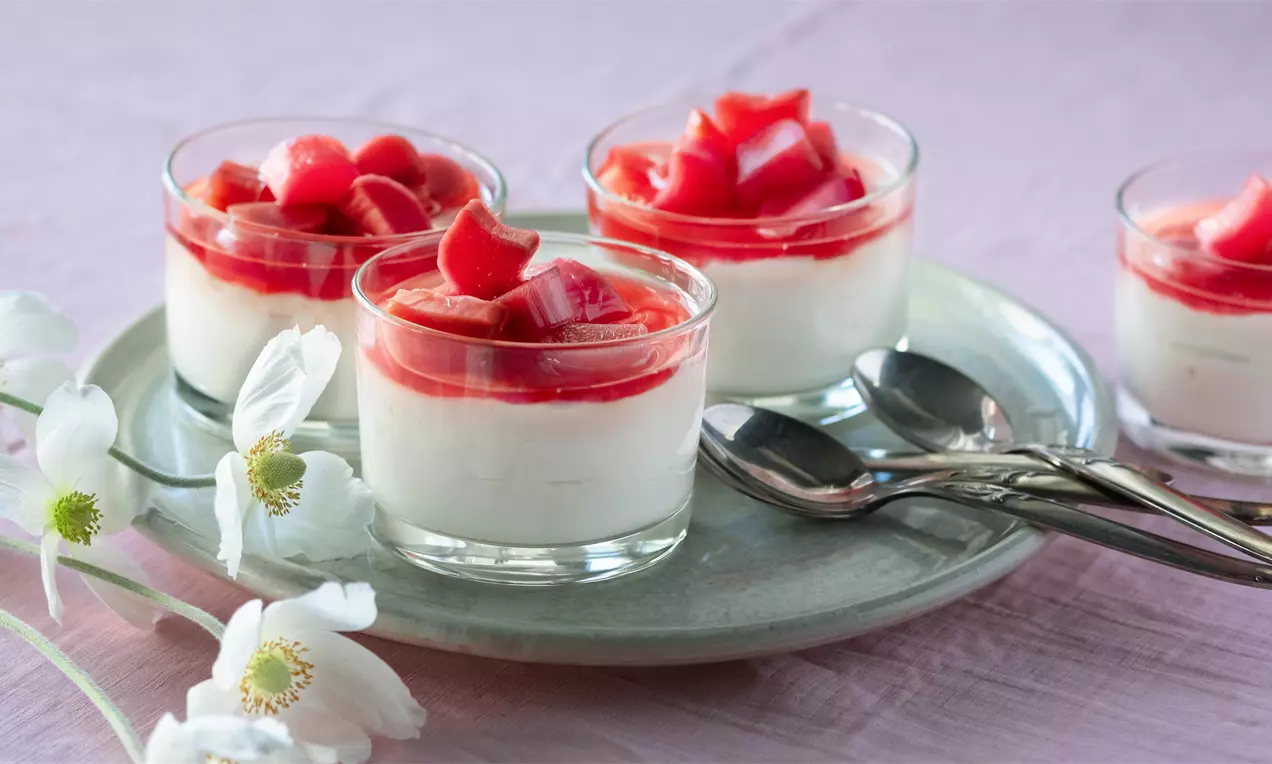Крем в стъклена чаша с бял слой основа, покрит с ярък червен топинг и парченца розов ревен, сервирано върху плато до цветя и лъжици