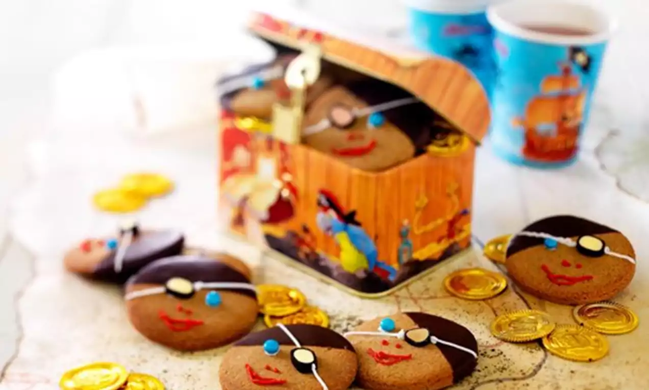 Ronde piratenkoekjes met bruine hoedjes van chocolade, gekleurde details en lachende gezichten, rondom een open schatkist en goudkleurige munten