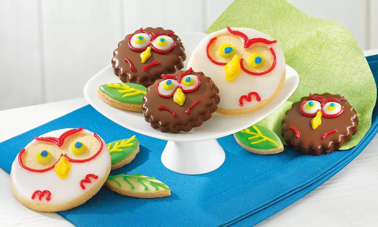 Biscuits hiboux ronds en sucre blanc ou chocolat avec yeux colorés et bec jaune sur une assiette blanche, posés sur un tissu bleu