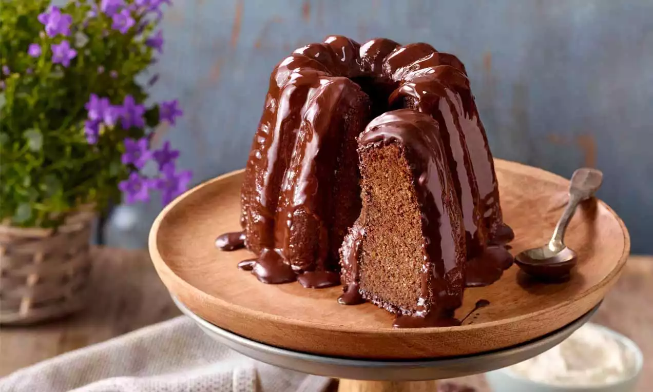 Dunkelbrauner Gugelhupf mit glänzender Schokoladenglasur und angeschnittenem Stück auf einem Holz-Teller neben einem kleinen Löffel