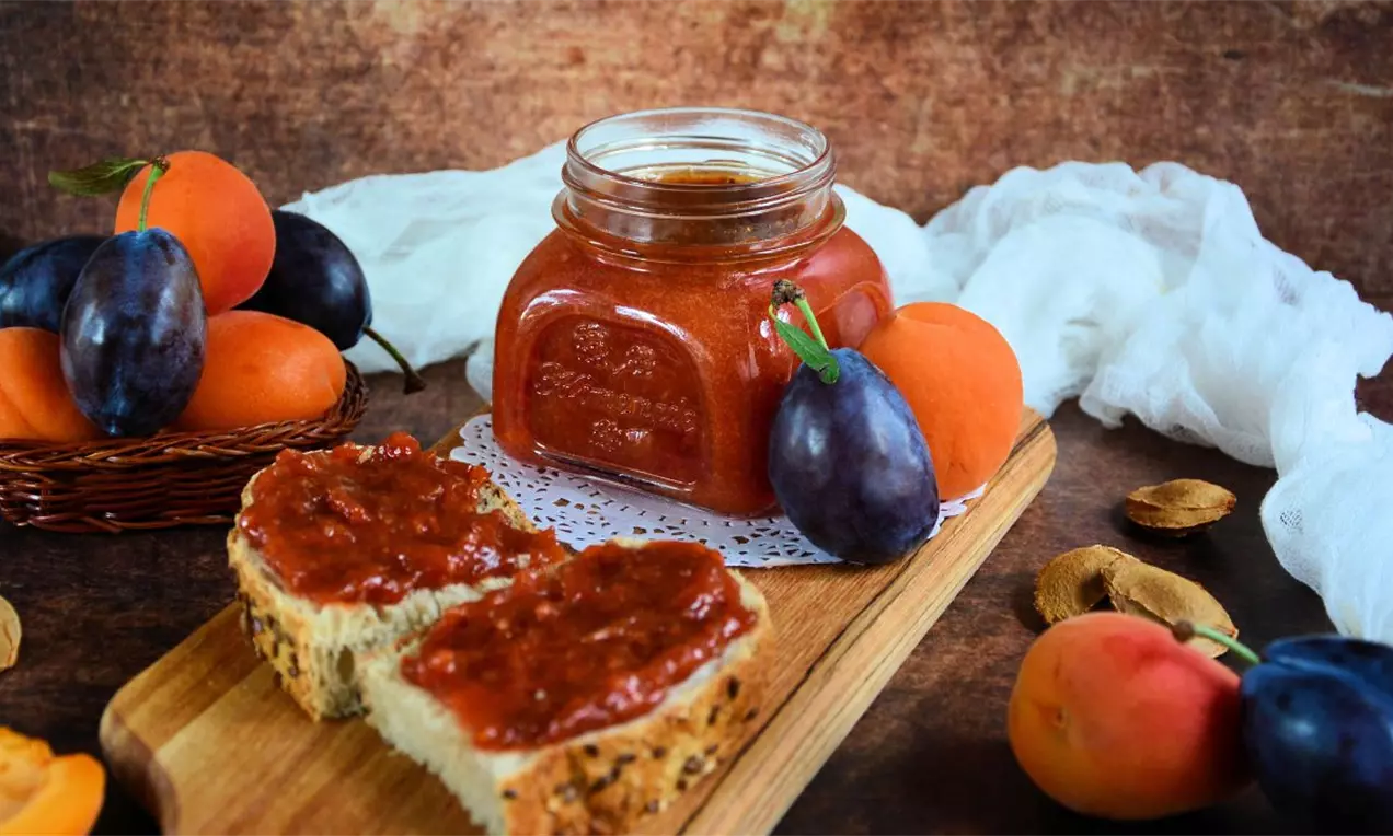 Staklena tegla punjena marmeladom jarke narandžaste boje okružena svežim šljivama i kajsijama, sa namazom na kriškama hleba