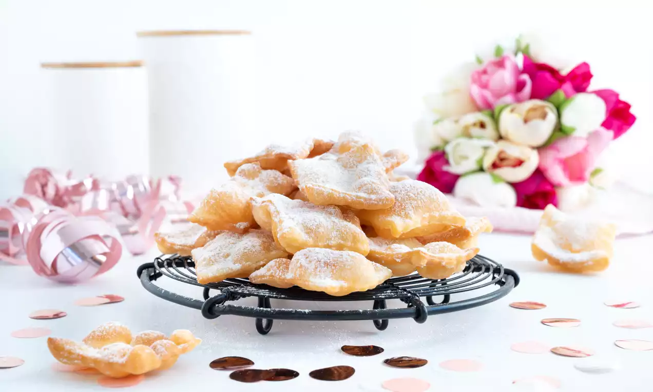 Goldbraune, knusprig-dünne Fasnachts-Chüechli unregelmäßiger Form, mit Puderzucker bestäubt auf schwarzem Gitter, umgeben von roséfarbener Dekoration