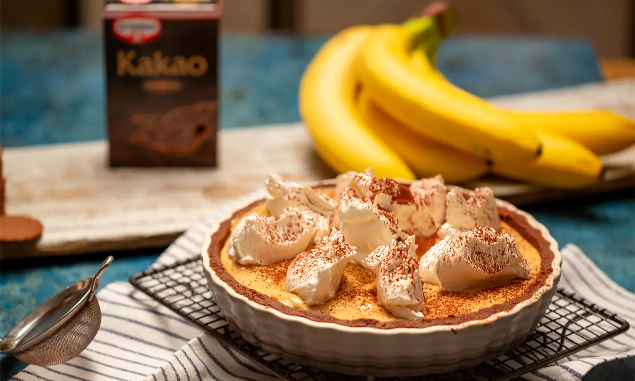 Банофи пай с кремообразен пълнеж, покрит със сметанови облаци и поръсен с какао, върху отразяваща златисто и кафяво метална решетка