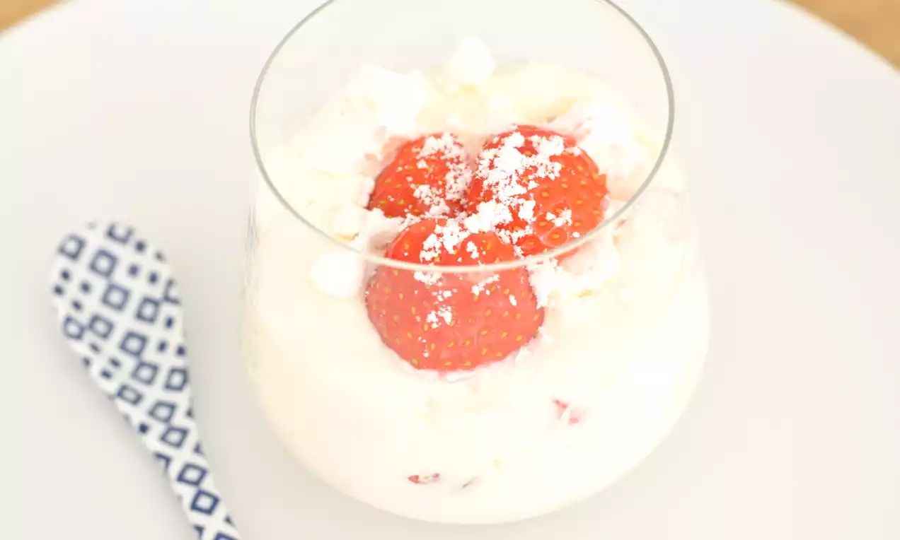 Glas met romige witte yoghurt, helderrode aardbeien en verkruimelde witte meringue op een wit bord met een lepel met blauw-wit patroon