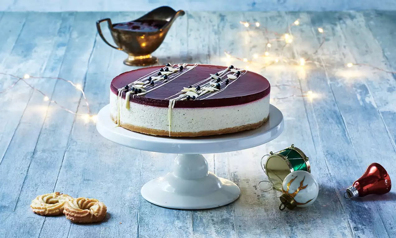 Rund ris à l'amande-kake med kremhvit ostefyll, brun kjeksbunn, mørkerødt gelélag med hvit sjokoladedekor på hvitt serveringsfat