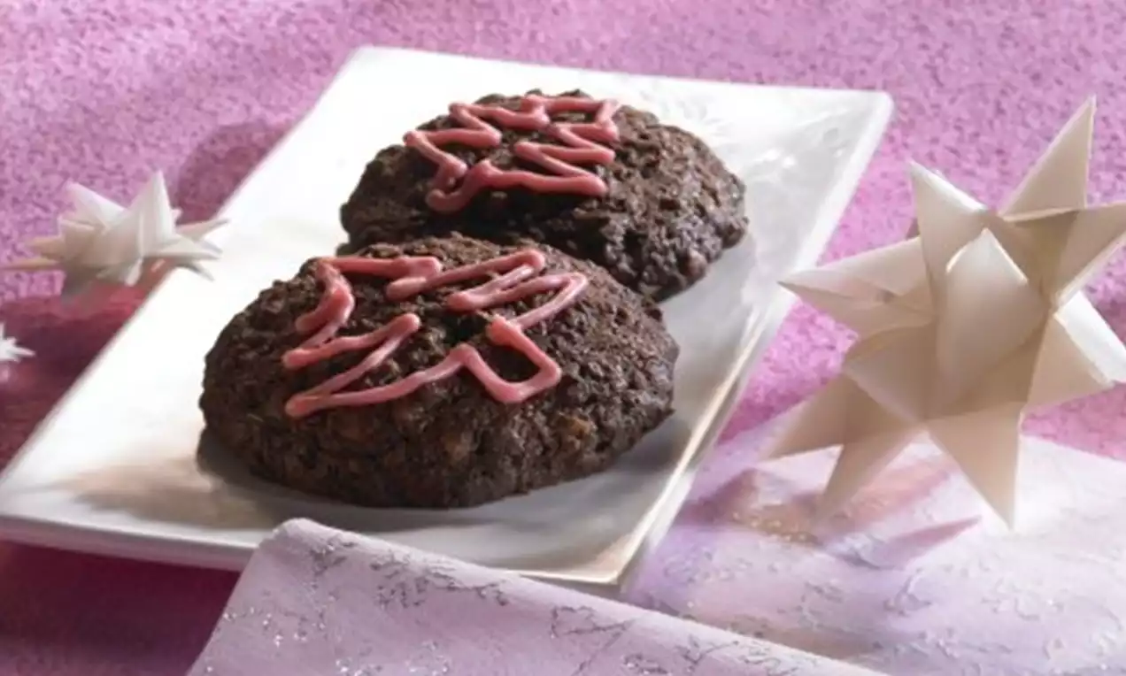 Two dark brown, round chocolate cookies with a crunchy texture and pink icing drizzled on top, served on a white plate against pink fabric