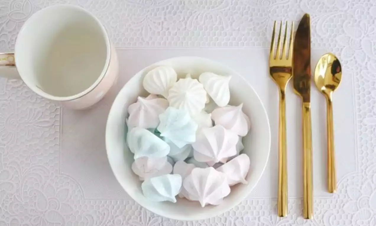 Leicht pastellfarbene Aquafaba-Baisers in zarten Rosatönen und Weiß sowie Hellblau in einer weißen Schale auf filigran gemustertem Tischset