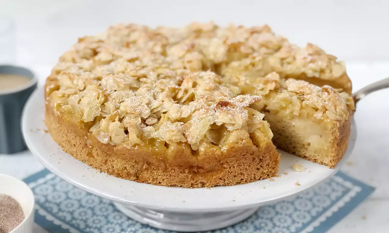 Veganer Apfelkuchen mit goldbrauner Mandel-Knusperhaube, saftiger Füllung und einer herausgelösten Ecke auf weißer Tortenplatte