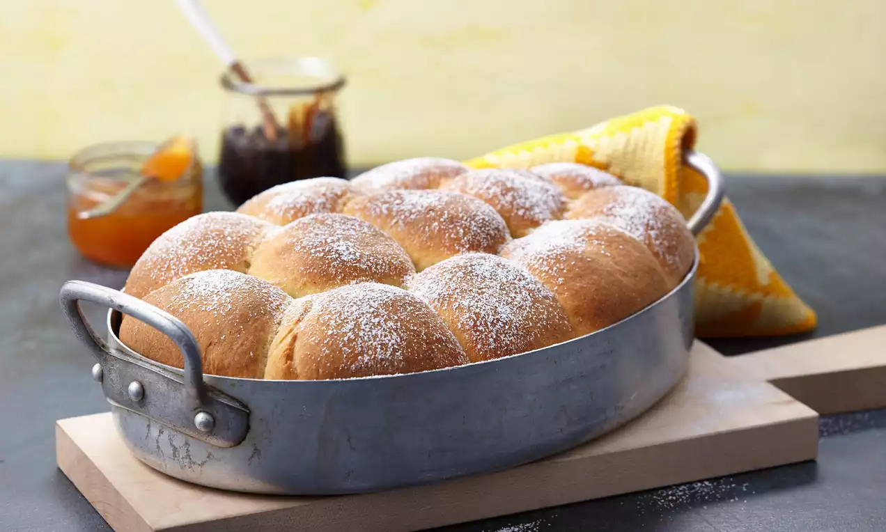Goldbraune, weich gebackene Buchteln mit Puderzucker bestäubt in einer ovalen Metallform, dahinter Marmeladegläser und ein gelbes Tuch