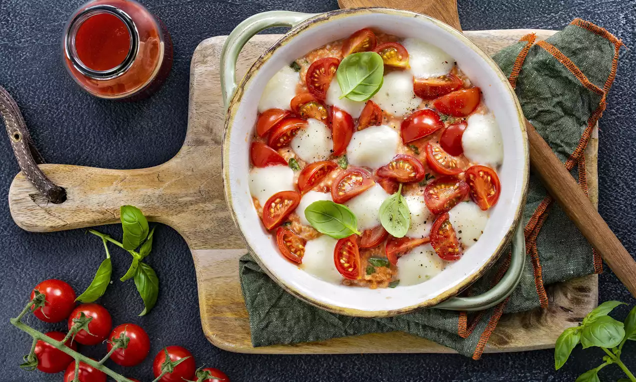 Tomaten-Mozzarella-Porridge mit weißer Mozzarella und roten Tomatenstücken, garniert mit grünen Basilikumblättern in einer runden weißen Schale