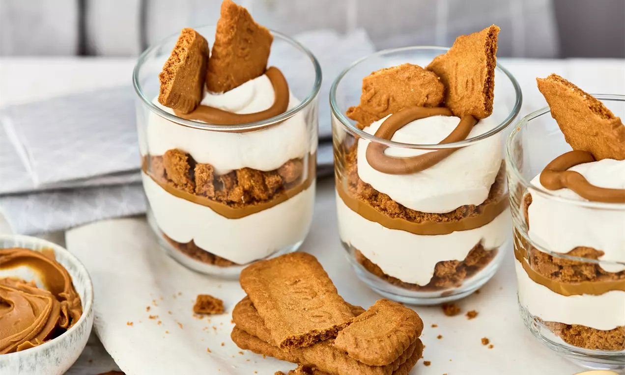 Veganes Lotus-Dessert in drei Gläsern, geschichtet mit Keksstücken, Creme und Lotus-Aufstrich. Garniert mit ganzen Keksen. Daneben goldene Löffel, extra Kekse und eine Schale mit Aufstrich.