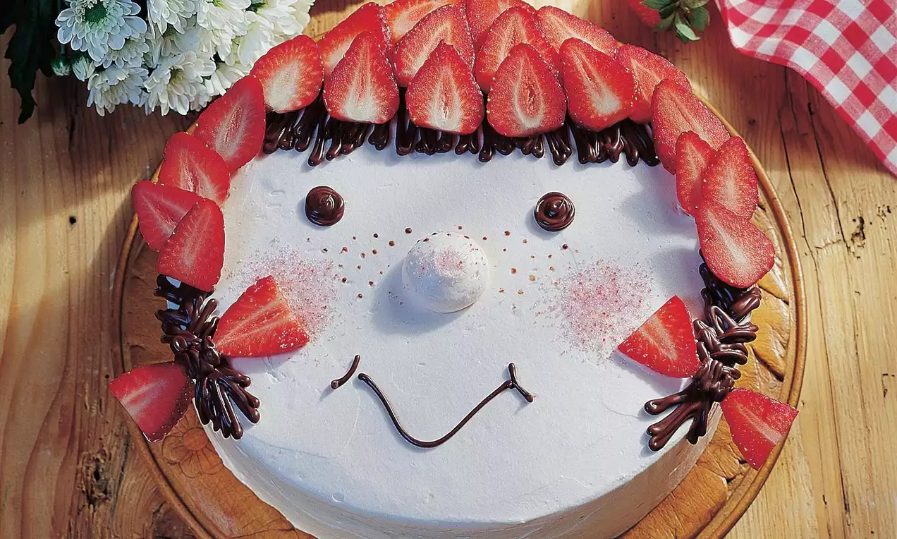 Tort ozdobiony białym kremem, truskawkami ułożonymi w kształt włosów, czekoladowymi dekoracjami i cukrowym noskiem na drewnianej paterze