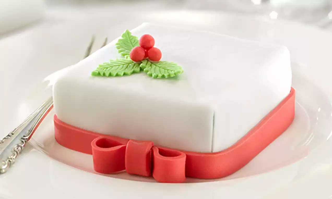 Mini tarta cuadrada cubierta de fondant blanco con cinta roja decorativa, hojas verdes y frutos rojos sobre un plato blanco