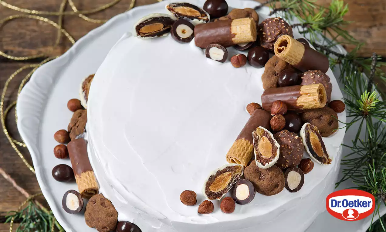 Ořechový dort s hladkým bílým krémem, dekorovaný čokoládovými tyčinkami, lískovými oříšky, sušenkami a pralinkami na světlém talíři