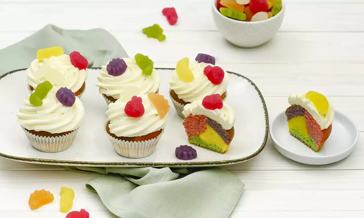 Papageienmuffins mit bunter, marmorierter Füllung, weißer Cremetopping-Spirale und bunten Gummitieren auf Teller und grünem Stoff drapiert