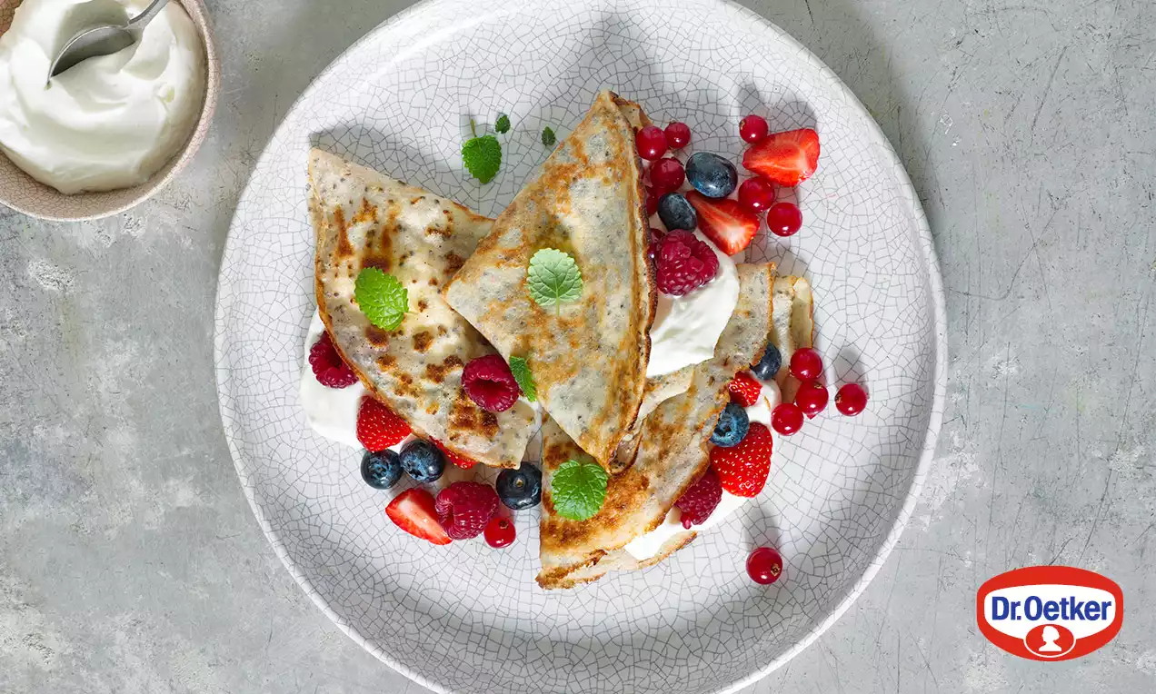 Zlatavé palačinky se světlou strukturou, složené na talíři s čerstvými červenými a modrými bobulemi, lžící bílého jogurtu a lístky máty