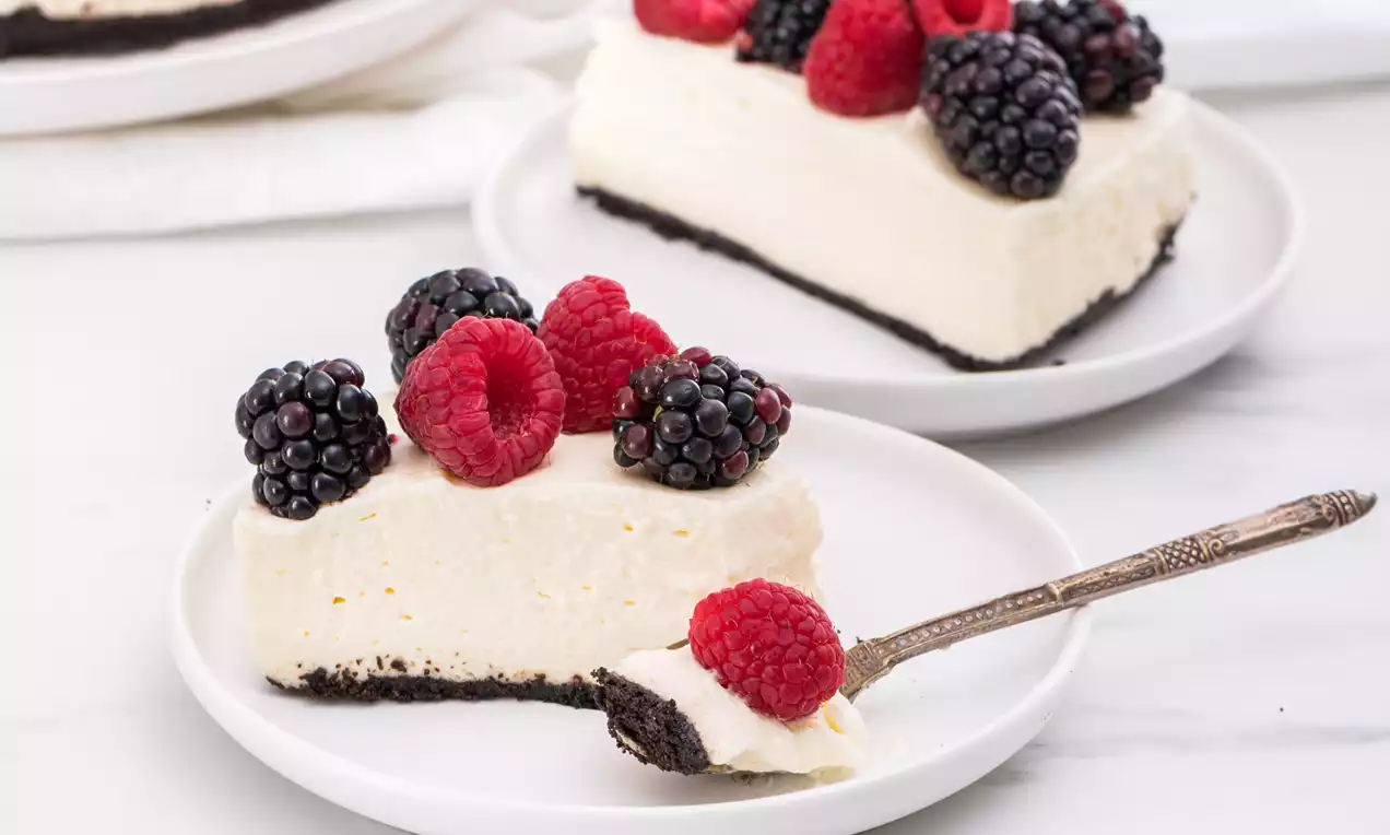 Tarta de queso cremosa sobre base oscura con frambuesas y moras brillantes dispuestas en la superficie sobre un plato blanco