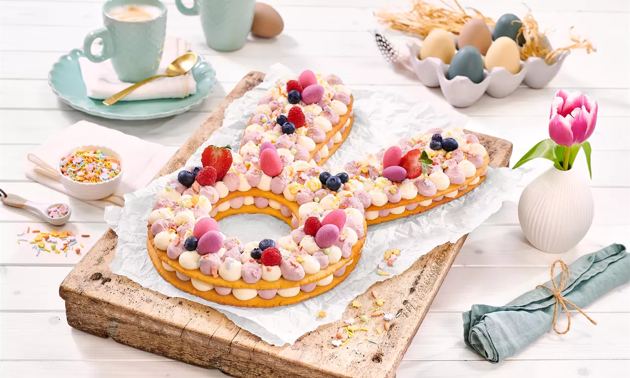 Torta v obliki črke z zlato rjavo podlago, prekrita s pisanimi kremnimi kupčki, jagodičevjem in dekoracijami na leseni podlagi.