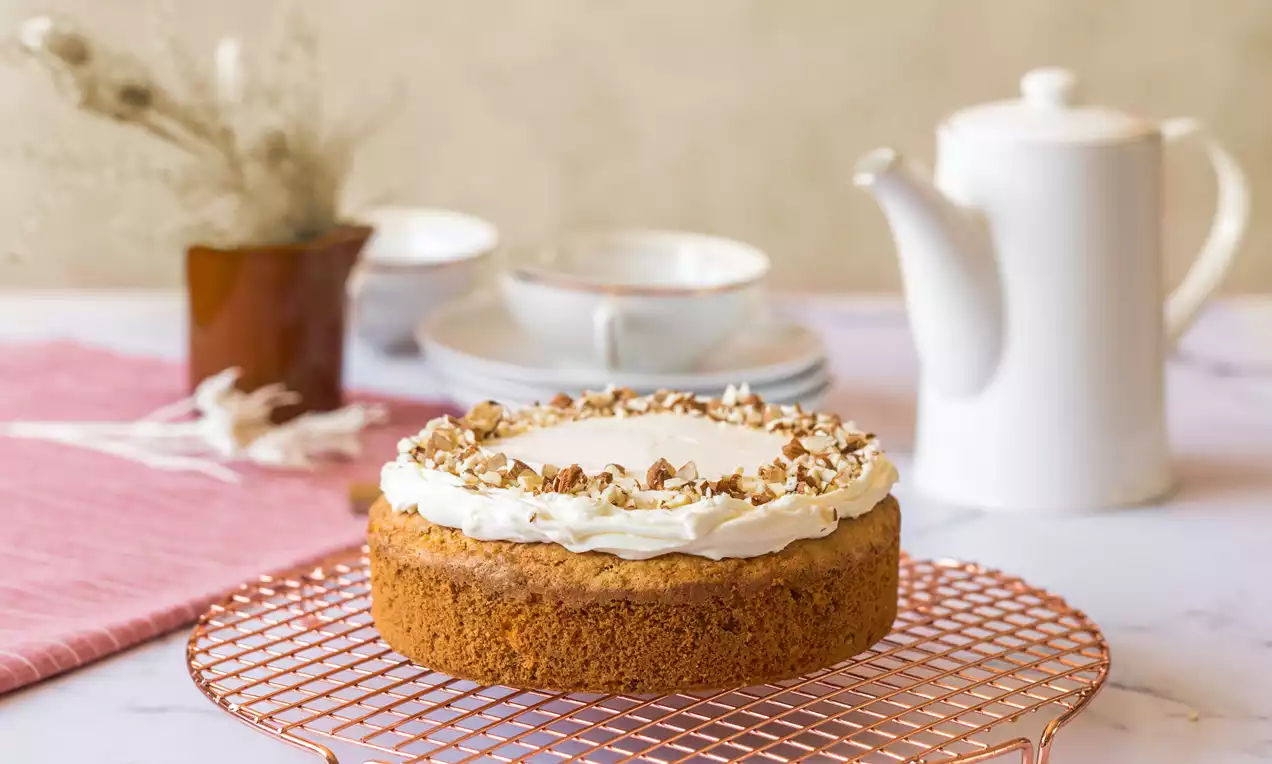 Bizcocho redondo de zanahoria de tono dorado con cobertura de crema blanca y nueces picadas sobre rejilla de cobre rosa