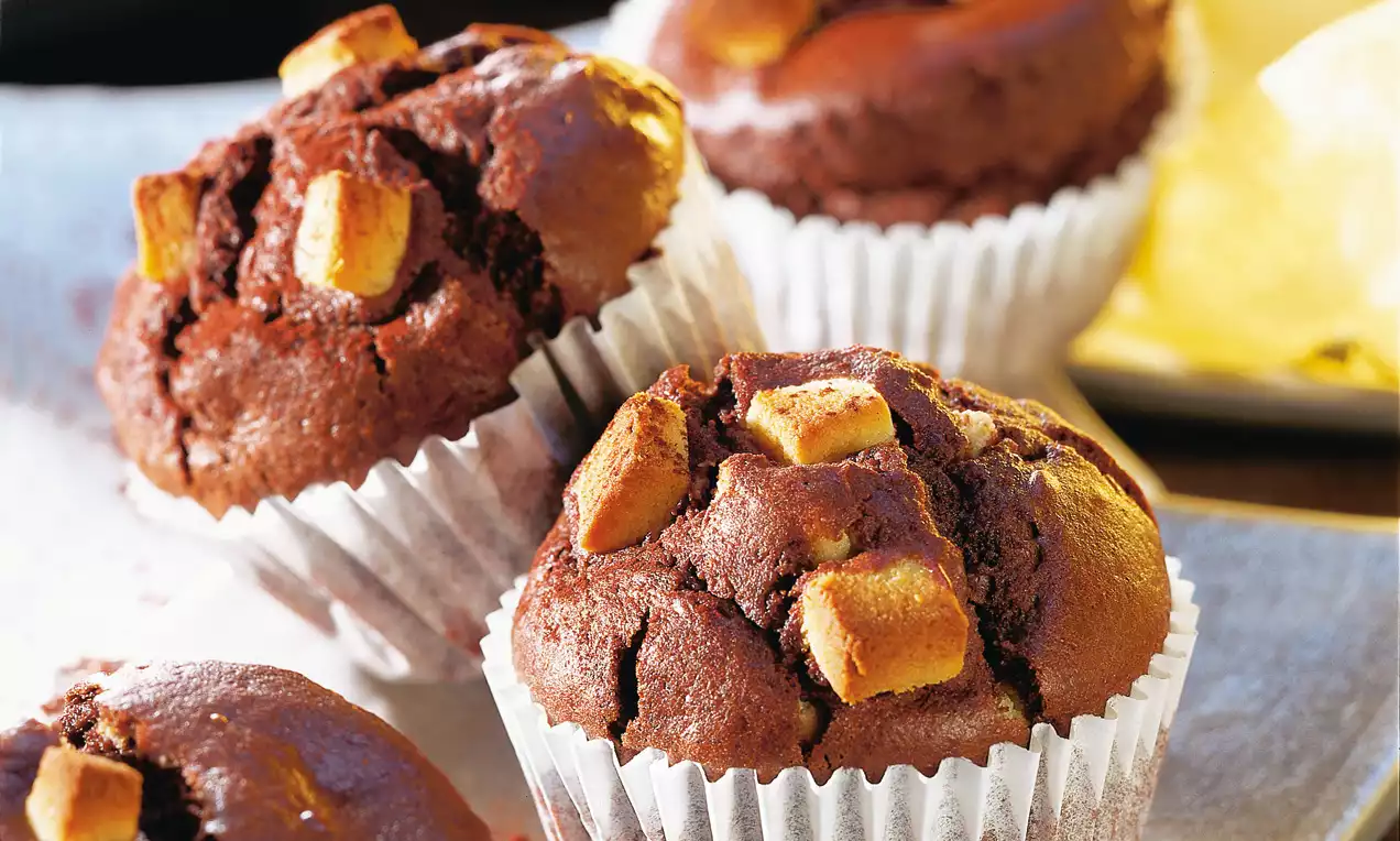 Muffins moelleux au chocolat brun foncé avec morceaux dorés de pâte d'amande, coiffés de plis blancs et disposés sur un plateau clair
