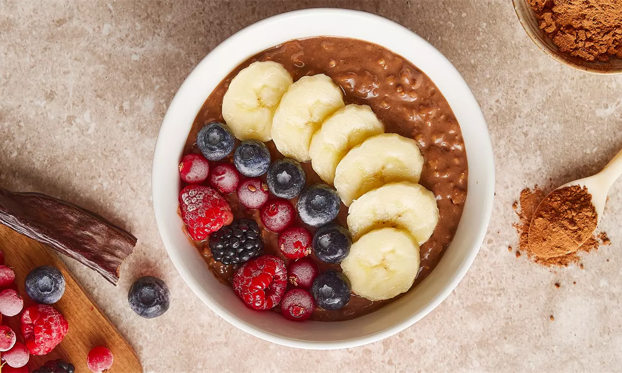 Kakaolu keçiboynuzlu yulaf lapası beyaz kasede, üzerine muz dilimleri ve karışık orman meyveleri eklenmiş, yanına kakao ve keçiboynuzu yerleştirilmiş