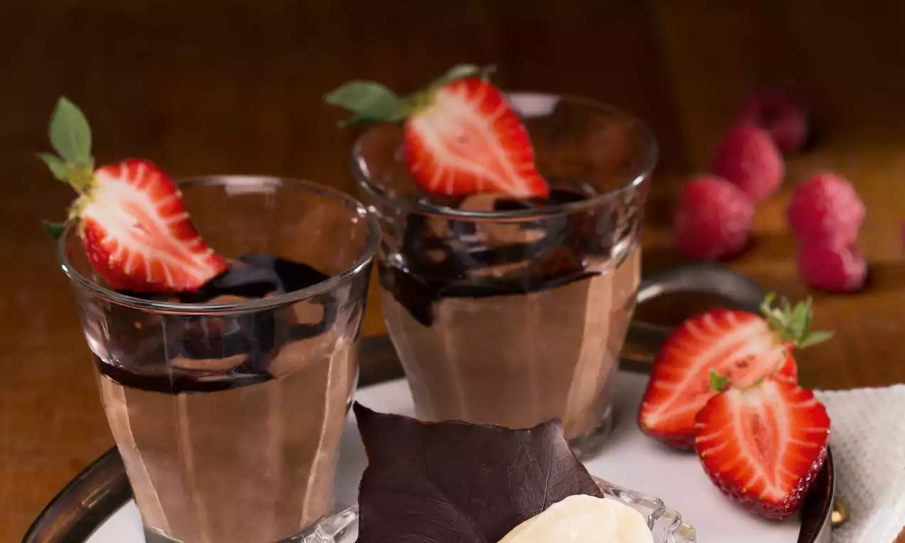 Zwei Gläser mit dunkler Schokomousse, garniert mit glänzendem Schokoladenspiegel und frischen halbierten Erdbeeren auf einem Tablett