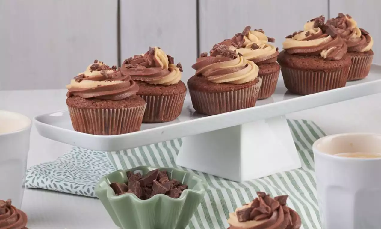 Schoko-Erdnuss-Cupcakes mit zweifarbigem Cremetopping, dekoriert mit Schokostückchen, auf weißer Etagere neben grüner Schale und Bechern